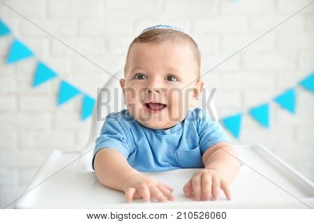 Cute baby in kippah sitting on high chair against brick wall