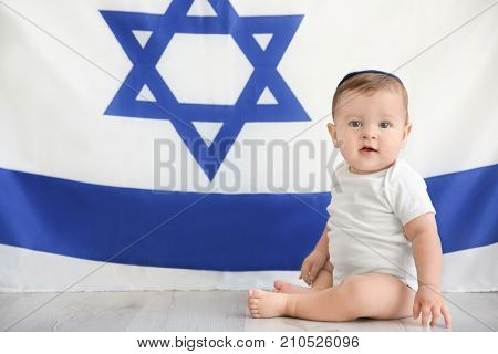 Cute baby in kippah sitting on floor against big flag of Israel