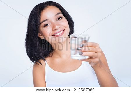 Staying hydrated. Pleasant cheerful woman in a white tank top holding a glass of water while standing isolated on a white background
