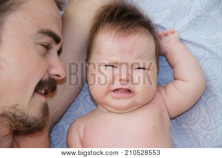 father and crying infant baby portrait lying on bed together exhausted parent