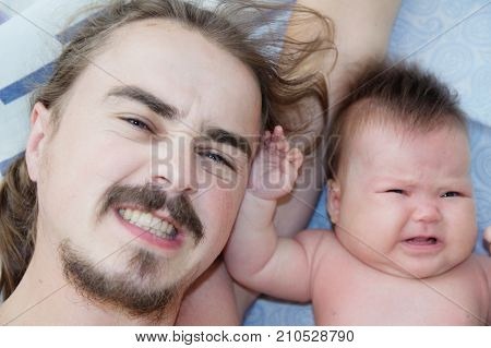 father and crying infant baby portrait lying on bed together exhausted parent