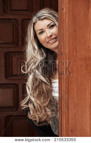 Beautiful girl peeks through the door peeking Out of Doorway. GIrl with low lighting a massive door covers half face closeup. Suspicious and attentive look.