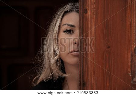 Beautiful girl peeks through the door peeking Out of Doorway. GIrl with low lighting a massive door covers half face closeup. Suspicious and attentive look.