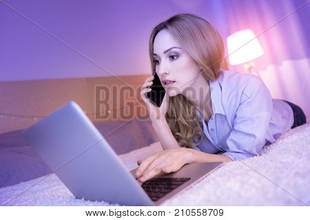 Discussing work. Attractive dark-eyed young woman talking on the phone and looking concentrated while working on her laptop