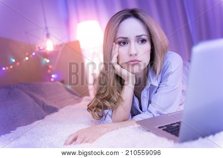 Waiting for news. Attractive dark-eyed bored woman lying on bed and looking at the laptop while waiting for something