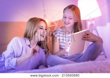 Being a parent. Cheerful loving blond mom and daughter smiling relaxing on the sofa while the girl showing something on her tablet