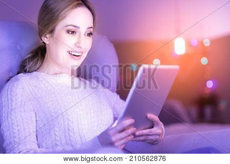 Pleasant news. Beautiful young blond woman emitting happiness and smiling while holding and looking at her tablet