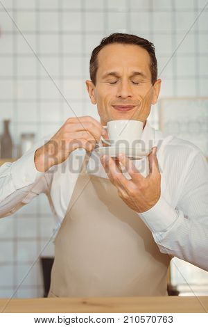 Wonderful smell. Happy delighted cheerful man holding a cup of coffee and smelling it while enjoying the smell