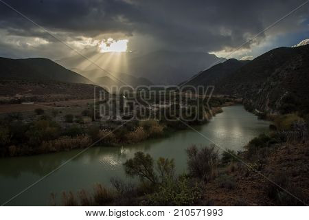 Shafts of sunlight shine through the clouds at sunset over a river in dry countryside