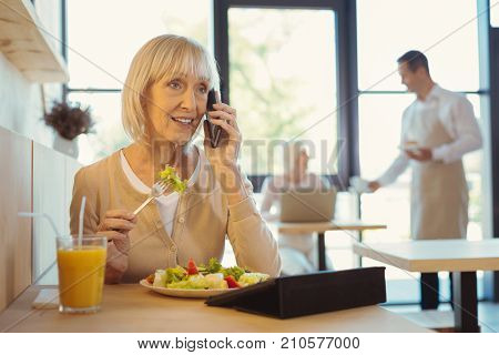 During lunch. Pleasant nice aged woman eating a salad and talking on the phone while having lunch