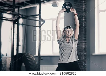 Gaining strength. Active senior woman standing in the gym and lifting up a weight disc with her both hands while performing muscle-strengthening exercises.