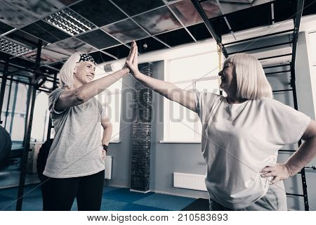 Great team. Cheerful senior women standing in the gym and giving each other a high five while smiling at each other pleasantly