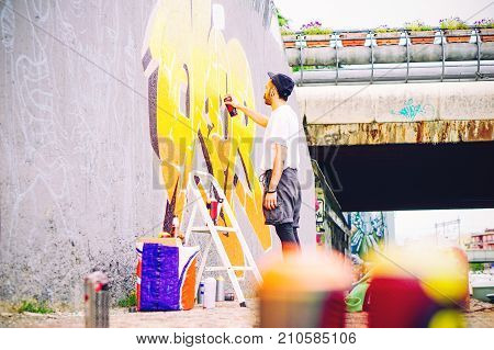 Street artist painting a colorful graffiti on a grey wall under bridge - Young graffiter writing and drawing murales with yellow spray - Urban lifestyle and modern art concept