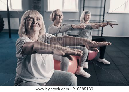 Workout with pleasure. Three charming lively senior women sitting on fitness balls, stretching out their hands and smiling pleasantly