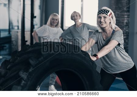 Role model. Pleasant senior female personal trainer showing a complex of workout exercises with a big tire to her clients standing in the background and paying much attention