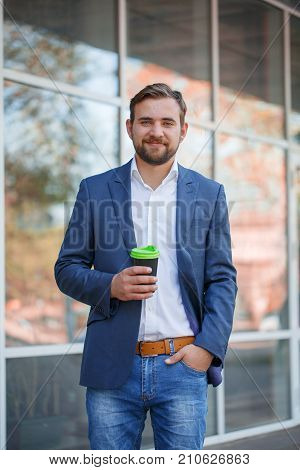 Attractive business man with beard drinking coffee and posing on the camera on the street. Panoramic wondows on the background. Close-up of man.