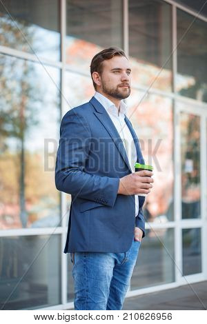 Attractive business man with beard drinking coffee and posing on the camera on the street. Panoramic wondows on the background. Close-up of man.