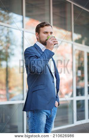 Attractive business man with beard drinking coffee and posing on the camera on the street. Panoramic wondows on the background. Close-up of man.