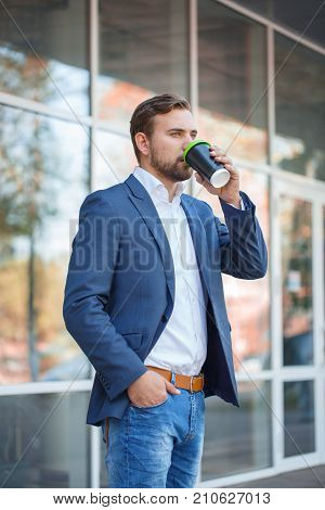 Attractive business man with beard drinking coffee and posing on the camera on the street. Panoramic wondows on the background. Close-up of man.