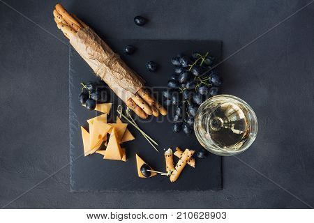 Cheese plate served with crackers grapes and glass of white wine on dark background. Old gouda cheese on tasting plate. Top view