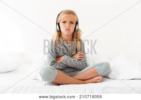 Upset female kid in headphones sitting with crossed arms and legs in bed, looking at camera