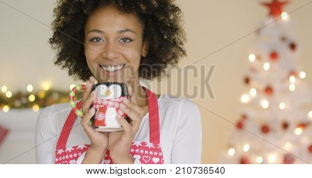 Pretty young housewife relaxing at Christmas standing in her festive apron enjoying a mug of coffee in front of the tree