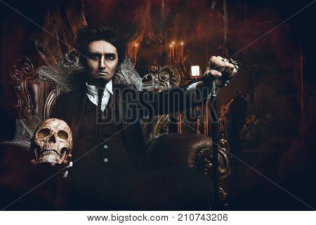 Halloween. Frightening gloomy man in a black tailcoat sits in an armchair with a skull in an old abandoned castle. The Dark Lord. Vampire man. 