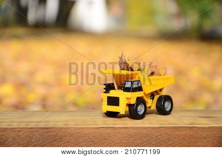 A Small Toy Yellow Truck Is Loaded With Yellow Fallen Leaves. The Car Stands On A Wooden Surface Aga