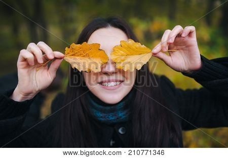 Young woman covered her eyes with yellow autumn leaves and cheerfully smiles. walking in the autumn park and fun with yellow autumn leaves.