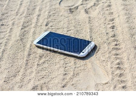 Lost white smartphone with crack on the screen lying in the sand