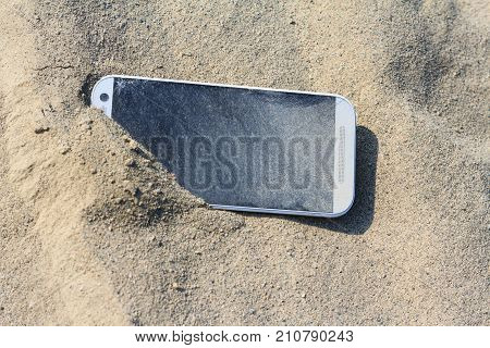 Lost white smartphone with crack on the screen lying in the sand