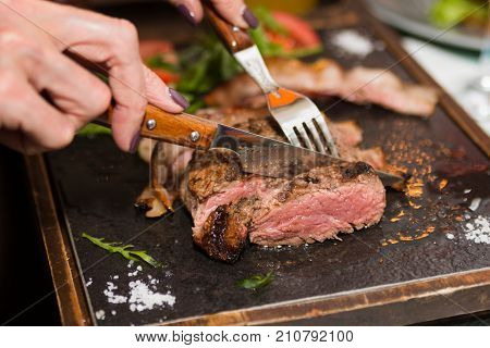 Woman hand holding knife and fork cutting grilled beef steak on stoned plate. Selective focus