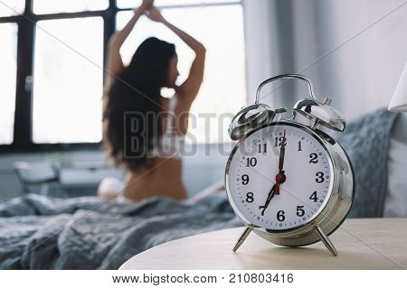 Amazing brunette is sitting on the grey bed in a big light room and stretching. The alarm on the table shows 7 a.m. so it's time to wake up and prepare for a new day. Close up