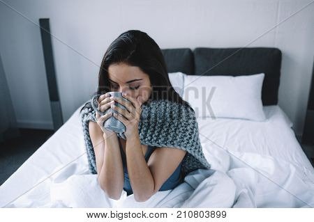 This girl is sitting on the bed and drinking some hot tea from the cup. She got sick this morning and wants to recover as soon as possible. Young woman will take some medicine to feel better later this day. Close up.