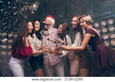 The bearded man in Santa's hat pours champagne into girl's glasses and smile. Modern Santa in Christmas hat have a fun with girls in the club with confetti. Happy New Year 2018