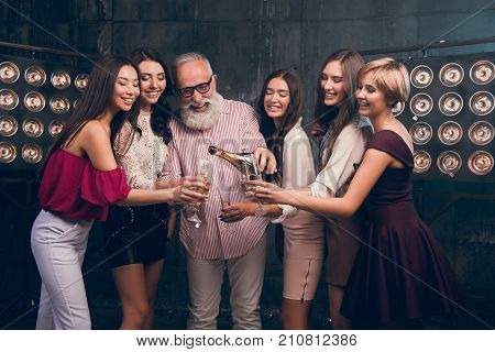 Modern Santa pours champagne into girl's glasses and smile. Happy Christmas day with fun group of attractive girls ad bearded man