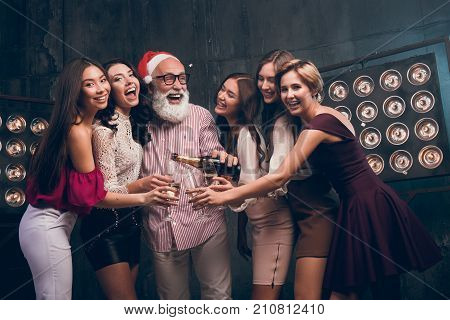 The bearded man in Santa's hat pours champagne into girl's glasses and smile. Modern Santa in Christmas hat have a fun with girls in the club. Happy New Year 2018