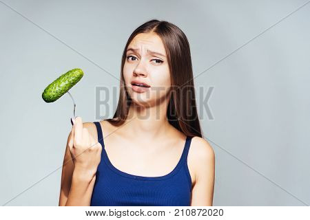 a young beautiful girl in a blue top wants to lose weight, holds a useful cucumber in her hands and looks perturbed