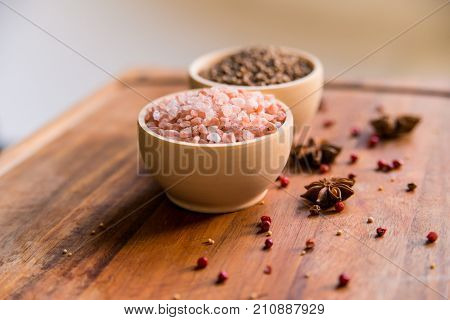 Pink Himalayan salt and coriander seeds in white bowls on wooden surface covered with anise and red peppercorn