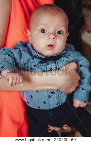 Portrait of newborn baby on mothers hands. Wearing blue shirt, tie bow and trousers. Lifestyle close up photo.
