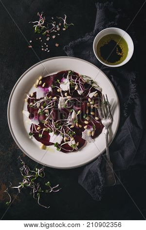 Vegetarian beetroot carpaccio salads with olive oil, goat cheese, pine nuts and sprouts in white plate on textile napkin over dark rusty metal background. Dark rustic style, healthy eating