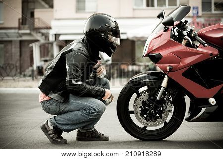 a guy a motorcyclist in a helmet and a leather jacket and jeans sits opposite a motorcycle of a sports red color and look at him face to face