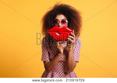Close-up portrait of playful african woman in sunglasses holding big red lips in front of her face, looking at camera, isolated on yellow background