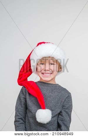 Little blond emotional boy in a festive red hat in a gray jacket. Little Santa Claus on a gray background.