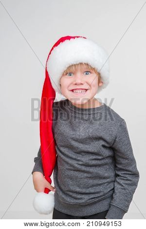 Little blond emotional boy in a festive red hat in a gray jacket. Little Santa Claus on a gray background.