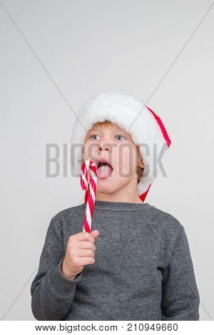 Little blond emotional boy in a festive red hat in a gray sweatshirt with a sweet stick. Little Santa Claus with lollipop.