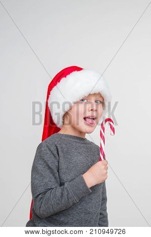 Little blond emotional boy in a festive red hat in a gray sweatshirt with a sweet stick. Little Santa Claus with lollipop.