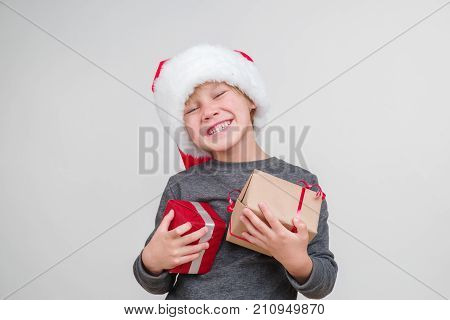 Little blond emotional boy in a festive red hat in a gray sweater with gifts. Little Santa Claus.