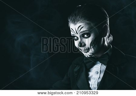 Portrait of a child boy with carnival make-up of a skeleton wearing elegant tail-coat. Studio shot over black background. Halloween. Dia de los muertos. The Day of the Dead.