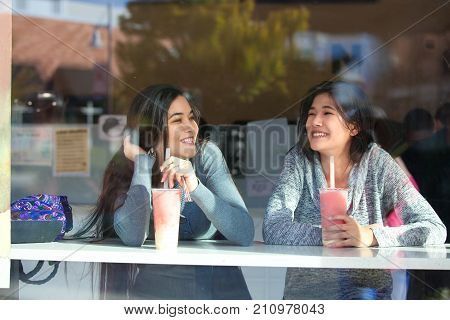 Two biracial teen girls or young women sitting together drinking boba tea inside cafe at counter